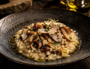 Prato de risotto de cogumelos silvestres com lascas de queijo e trufa, finalizado com um círculo de Azeite Invictus Premium dourado, realçando a textura cremosa.
